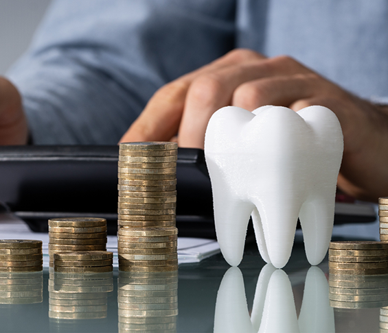 Person typing on a calculator on a desk next to a model of a tooth and several stacks of coins