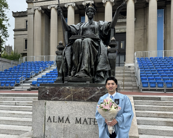 Doctor Choi in his graduation robes and holding a bouquet in front of a statue of a woman sitting in a chair with her arms held out